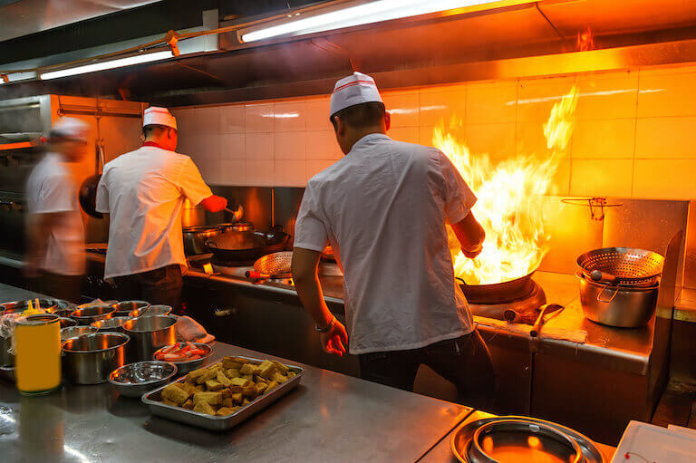 Two chefs stirring woks with a large flame