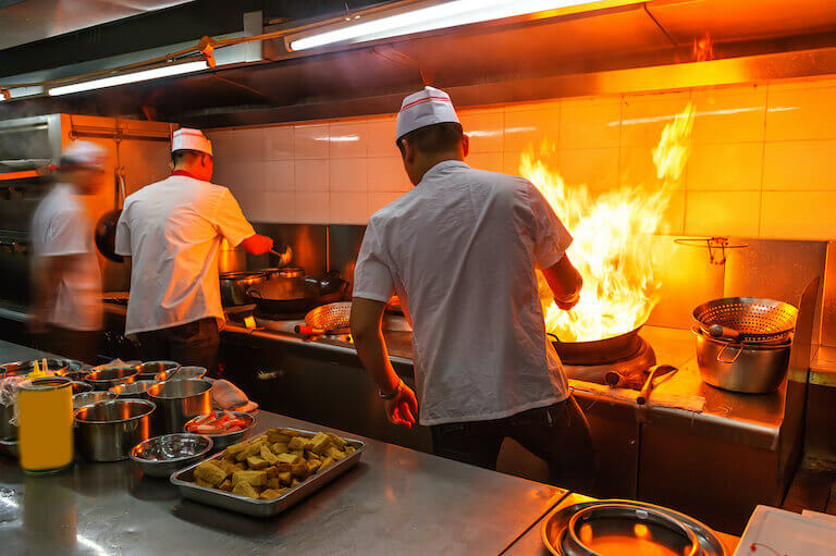 Three people stand at a professional stovetop, their backs to the camera. On the right, a person handles a large pan that has flames shooting up high.