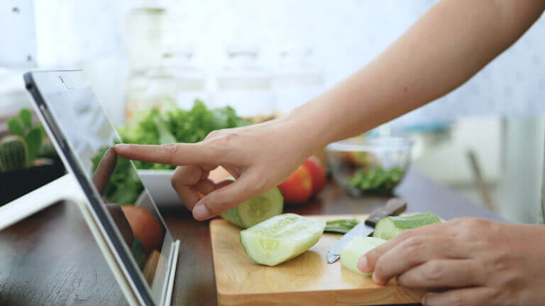 A chef taps the screen on a tablet while cutting cucumbers in the kitchen