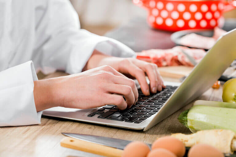 Chef typing on laptop computer on table with knife, eggs, and vegetables