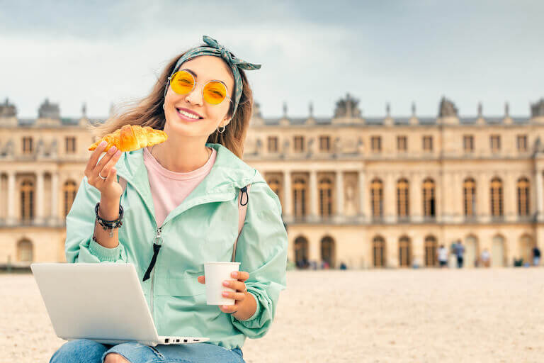 Young smiling woman sitting in front of The Louvre Palace in Paris, holding a croissant and coffee cup, with laptop in her lap
