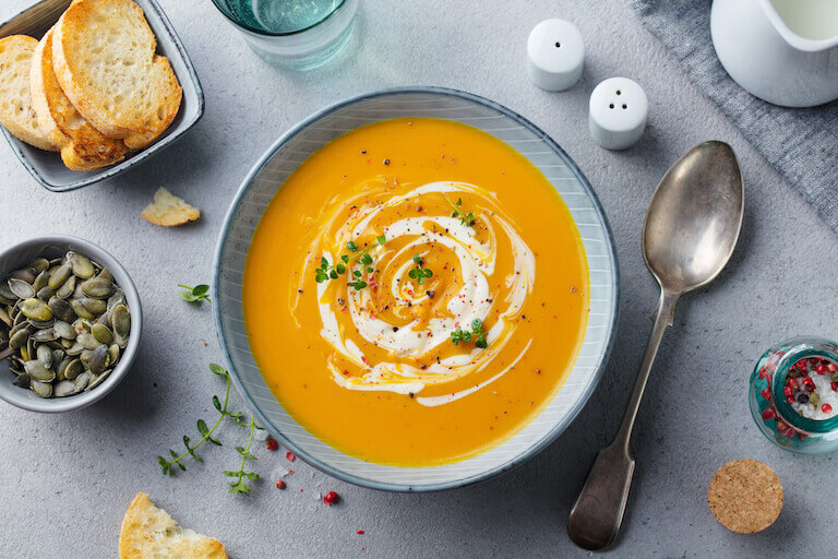 A bowl of pumpkin soup styled on a table with bread, seeds, salt, and a spoon