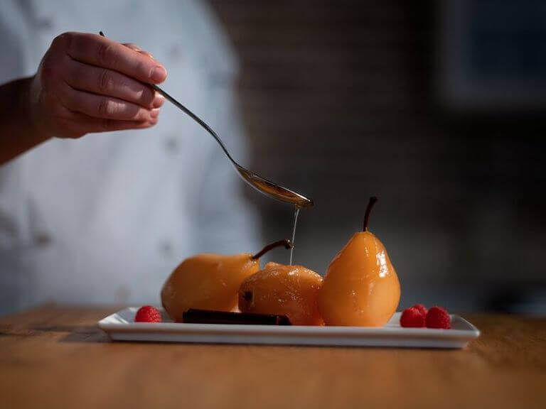 Close-up image of a chef using a spoon to drizzle glaze onto three peeled, poached pears, plated on a small rectangular dish alongside fresh raspberries and pieces of chocolate.