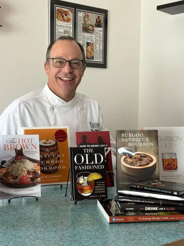 Escoffier Lead Chef Instructor Albert Schmid smiles in his chef's uniform, seated behind a display of several culinary and cocktail books he has authored.