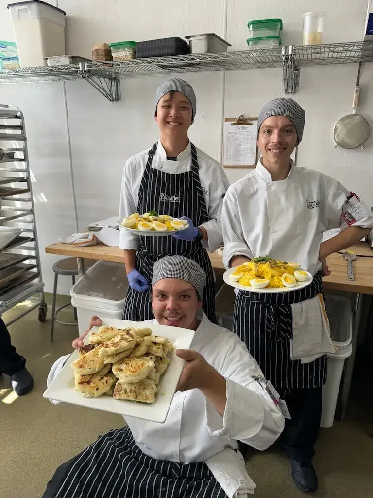 Three Escoffier culinary students smiling in a training kitchen while showcasing plates of prepared food, including sliced hard-boiled eggs and a platter of pan-seared flatbreads.