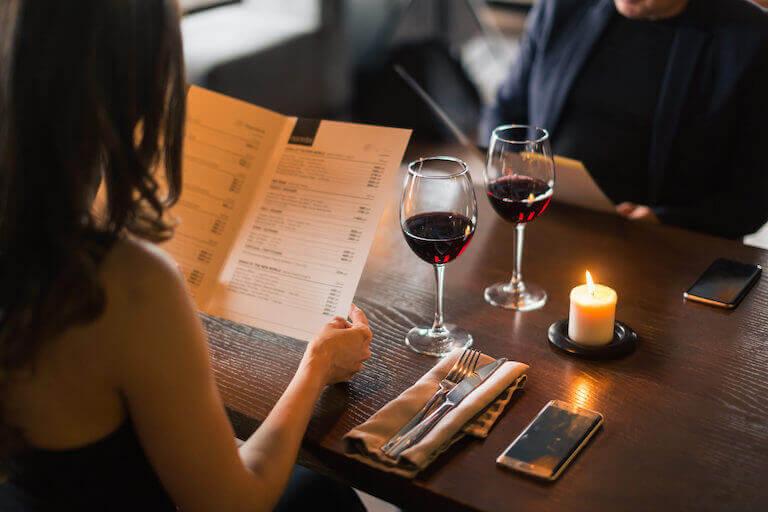 A man and a woman read menus while seated at a table with two glasses of red wine and votive candle.