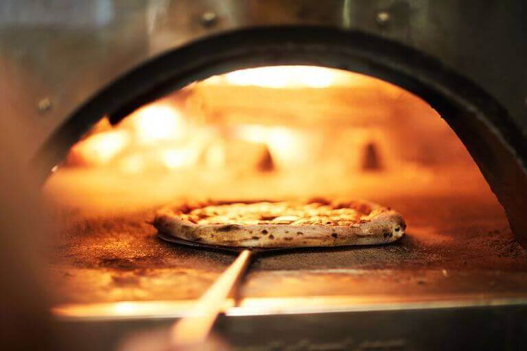 Close-up view of the inside of a wood-fired oven as a cheese pizza is removed using a pizza peel.