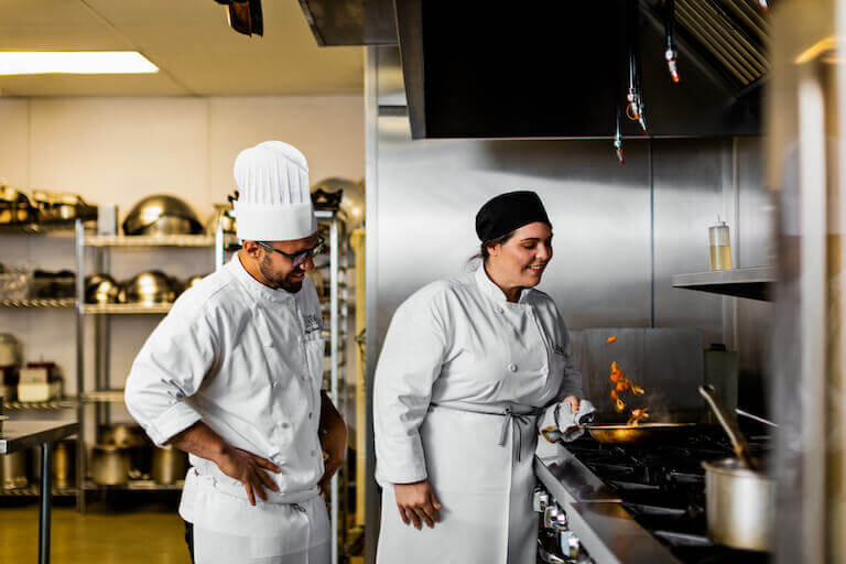 Escoffier student flipping ingredients in a pan while Chef Instructor watches