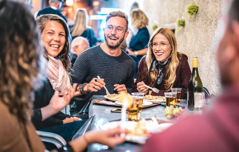 A group of smiling young people share a bottle of wine and light appetizers in a casual restaurant setting.