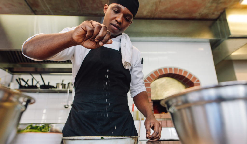 Chef sprinkling spices on dish in commercial kitchen.