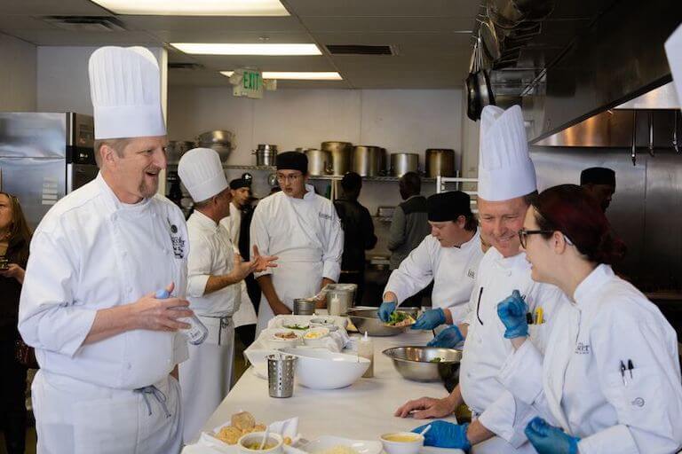 Chef instructor speaking with students as they assemble dishes in a kitchen