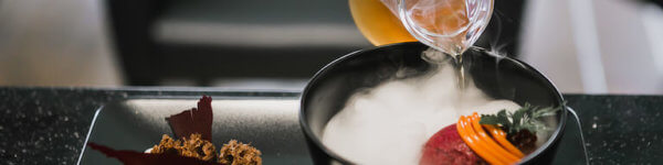 A chef pours liquid from a small glass pitcher into a black bowl, creating a thick cloud of vapor that spills over the edges, with crispy elements and garnishes artfully arranged on a sleek black plate.