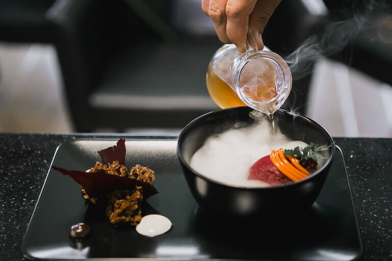 Chef pouring a sauce into a black bowl next to molecular food on a plate 