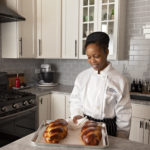 Female pastry student in home kitchen holding tray of baked bread