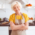 Happy older woman in apron preparing breakfast, crossed hands