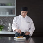 Male culinary student seasoning & garnishing a vegetable and meat dish in home kitchen