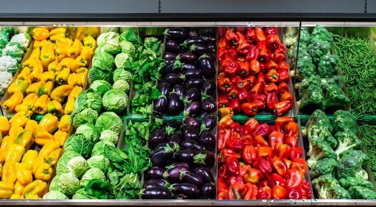 A grocery store display of yellow peppers, green cabbages, purple eggplants, red peppers, and green broccoli.
