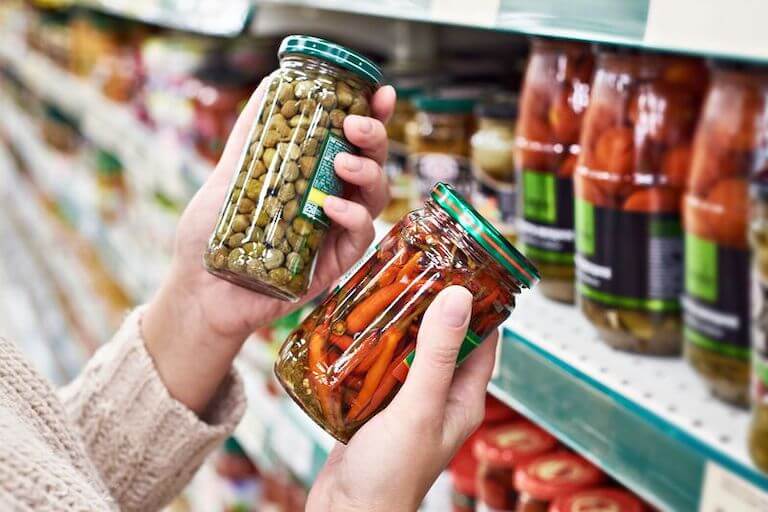 A person decides between two jars of pickled vegetables in a supermarket aisle.