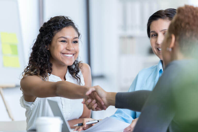 Two women shake hands in an office environment while a third looks on approvingly.