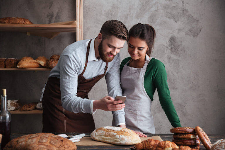 Two bakers stand in a rustic bakery surrounded by baked goods and use a smartphone to take a picture of a fresh loaf of bread.