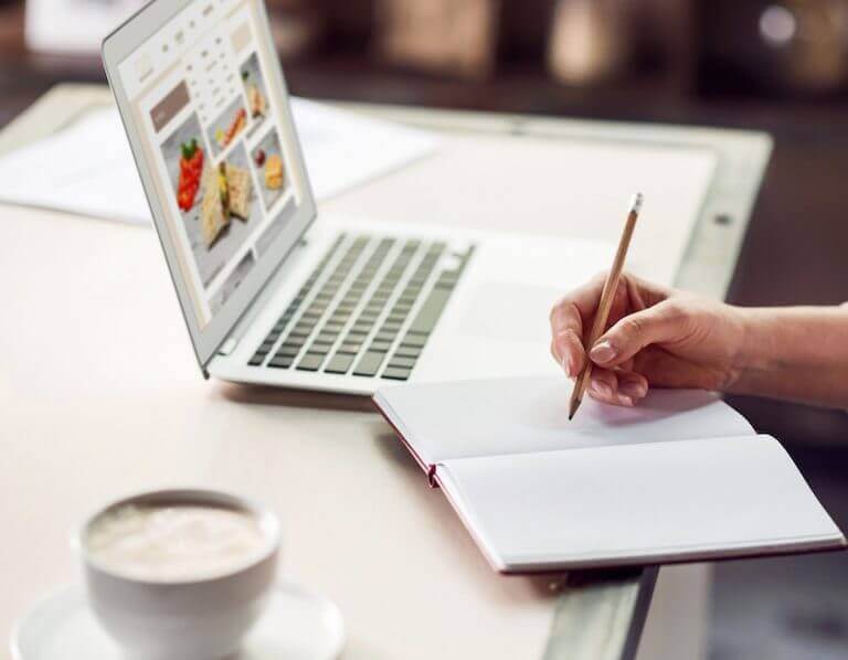 Student taking notes in journal while using laptop computer