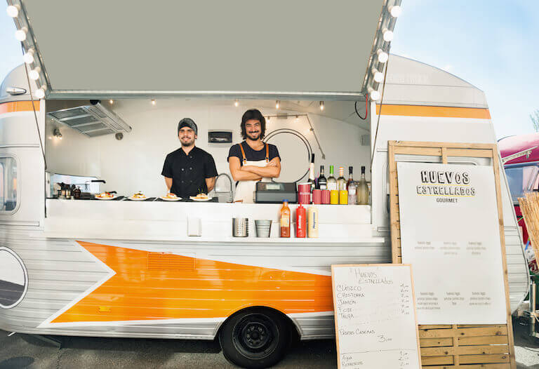 Two smiling chefs in an orange and white food truck with a menu for huevos estrellados