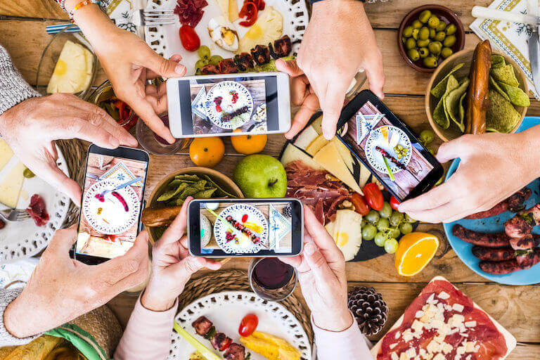 Four pairs of hands hold smartphone cameras over a wooden table covered in colorful, fresh foods.