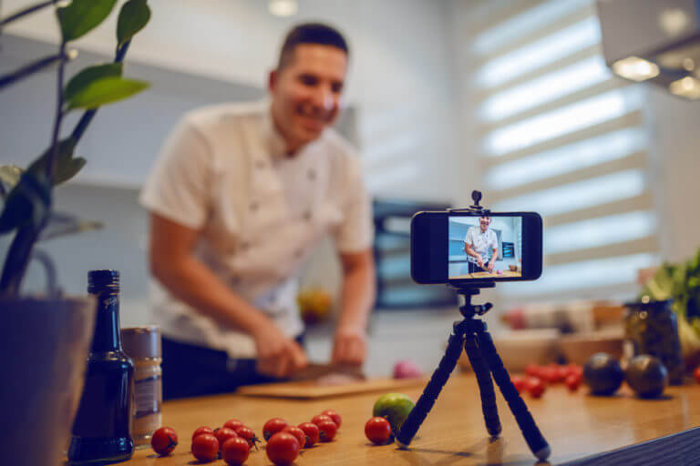 Smiling chef in uniform standing in kitchen and cutting onion while filming himself for blog