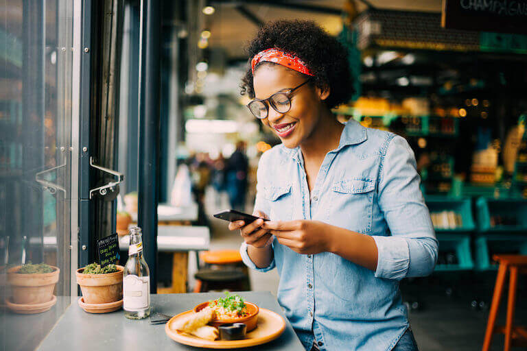 A woman in a denim shirt smiles while using her phone to photograph a plate of food by the window of an upscale cafe.