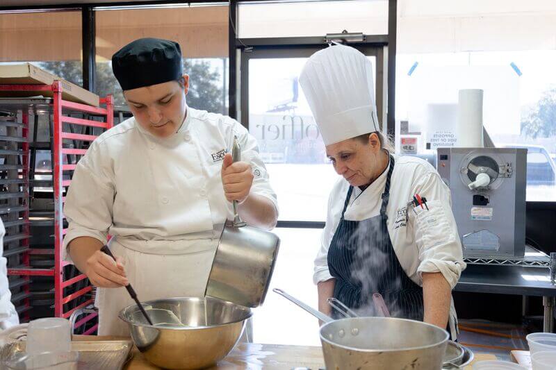 A culinary student pours ingredients into a large mixing bowl while a chef instructor stands nearby, watching.