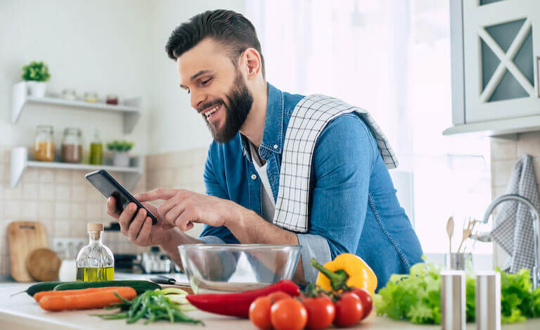 Bearded man using a smartphone while cooking with vegetables in the kitchen