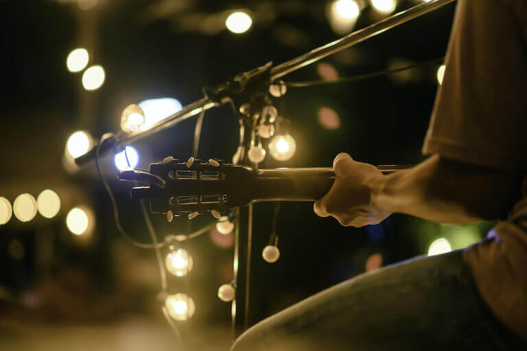 Close-up image of a person playing acoustic guitar and singing into a microphone in a dark space lit with string lights.