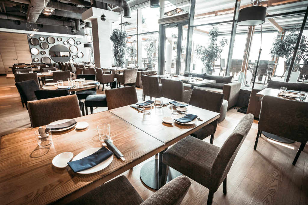 Contemporary restaurant with main hall with the wooden tables, chairs and velvet sofas, and glass wall with entrance to the terrace.