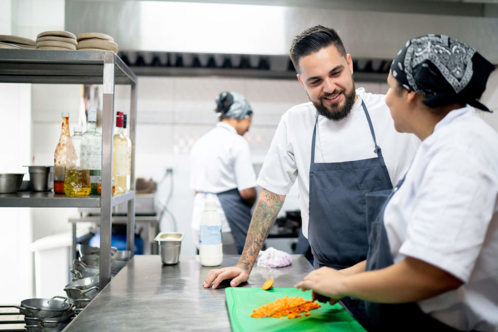 Female cook dicing carrotts while male chef with tattoos watches