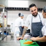 Female cook dicing carrotts while male chef with tattoos watches
