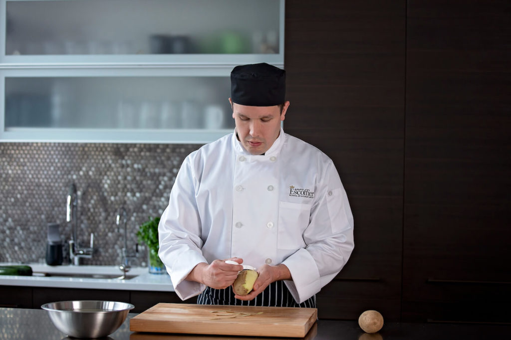 Escoffier online student peeling a potato with a peeler in home kitchen