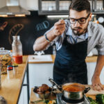 Male home cook with beard tasting soup from pot in kitchen