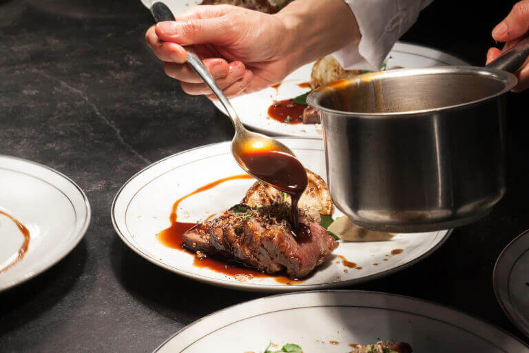 Pouring sauce using large spoon over a meat dish from metal pot