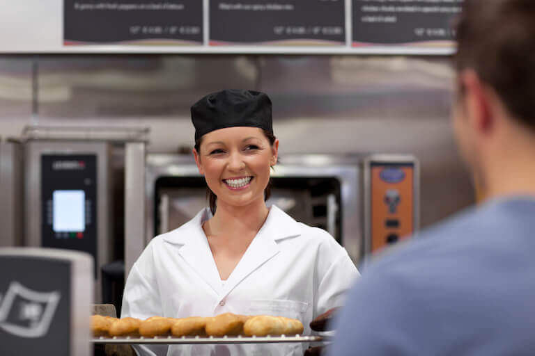 A person wearing a white chef coat and black chef’s hat holds a tray of baguettes and smiles at a person standing in the foreground with their back to the camera.