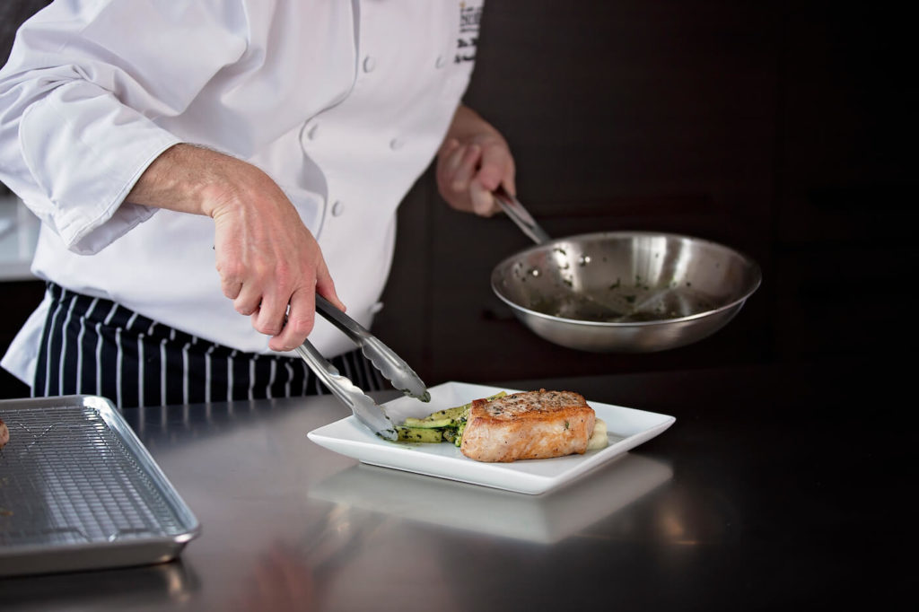 Chef using tongs to plate a meat and vegetable dish from metal pan