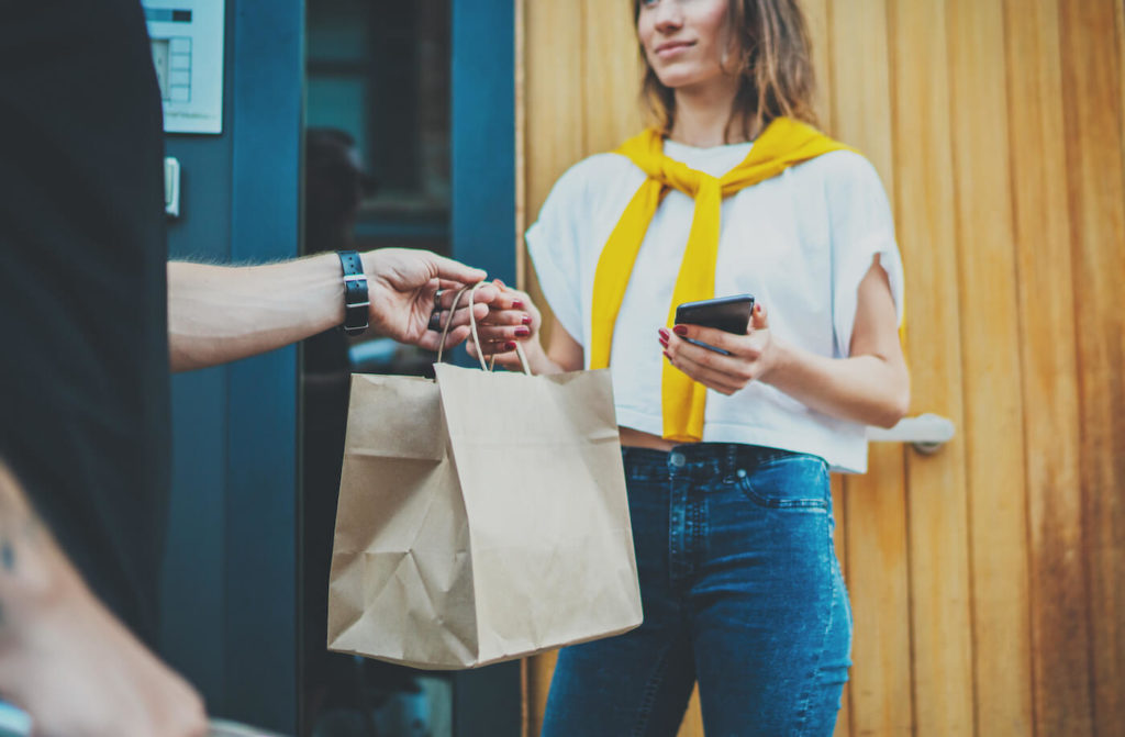 Male courier handing brown paper bag to woman with yellow sweater and phone for delivery food service at home