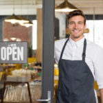 Smiling man with apron standing next to door of restaurant with open sign