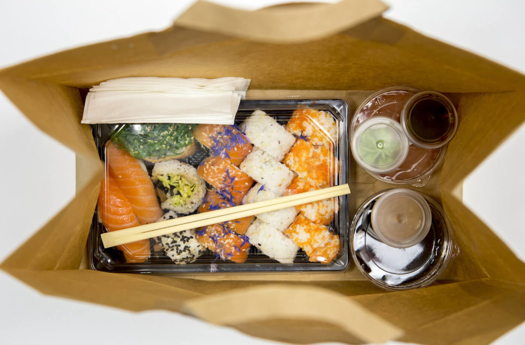 Sushi to go concept. Top view of takeaway box with sushi rolls and various sauce cups in brown paper bag