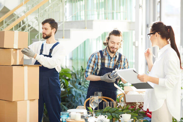 An event planner speaks with two movers delivering table decorations in a bright, airy event space.