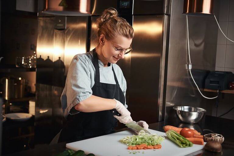 Smiling chef cutting carrots and other vegetables on a white cutting board in a kitchen