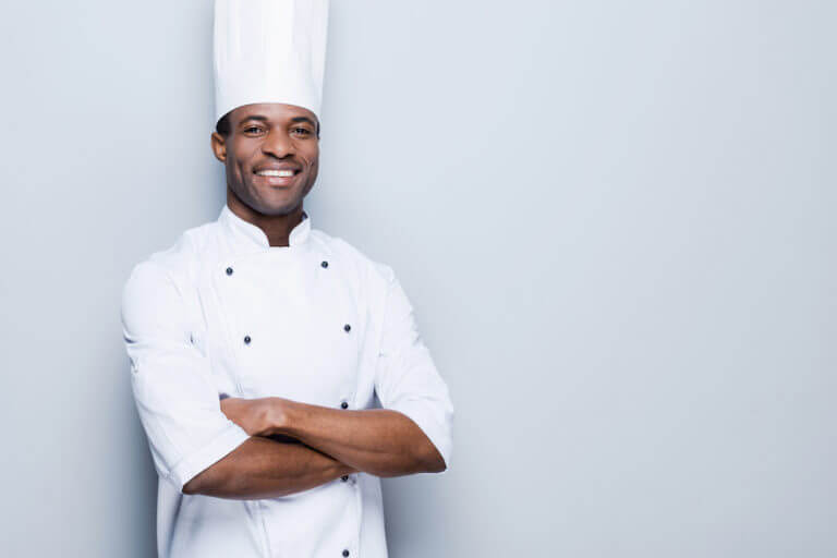 Smiling executive chef wearing a traditional tall white toque and white double-breasted jacket, arms crossed.