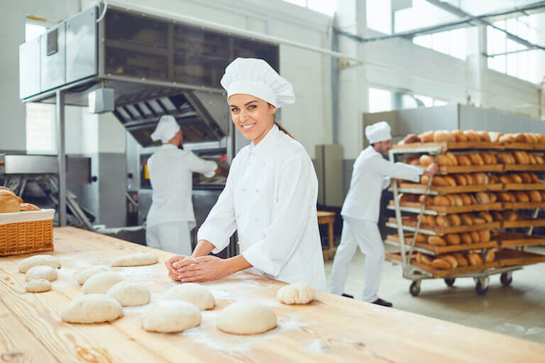 A baker in a white coat standing at a table shaping loaves of bread dough with bakers behind them at the oven and pushing a cart of bread.