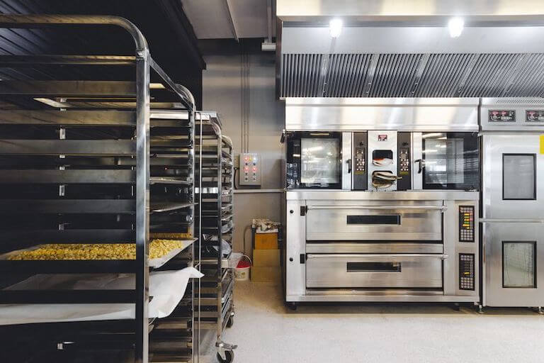 A rack of baked goods stands in front of industrial ovens in a professional kitchen.
