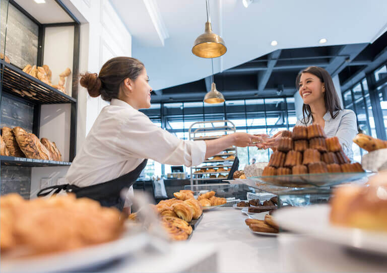 Bakery employee in a white shirt and black apron behind a counter lined with trays of baked goods hands a plate to a smiling customer on the other side of the counter.