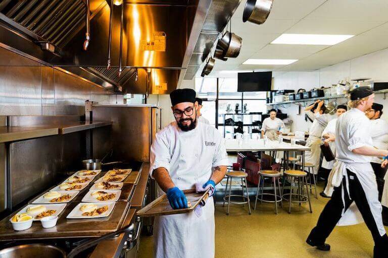 Escoffier student in chef uniform cleaning baking tray in kitchen with other students